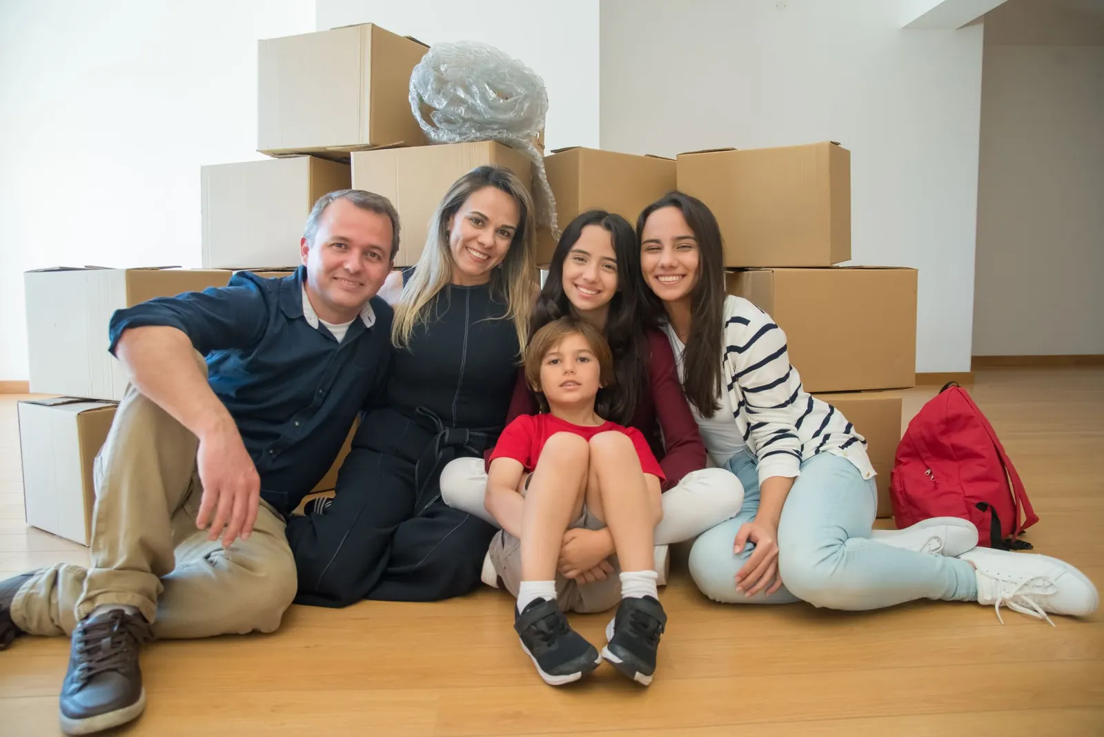 Smiling Latino-looking family of five (dad in navy shirt + khakis, mom in navy jumpsuit, two teen girls in burgundy/striped tops, young boy 
