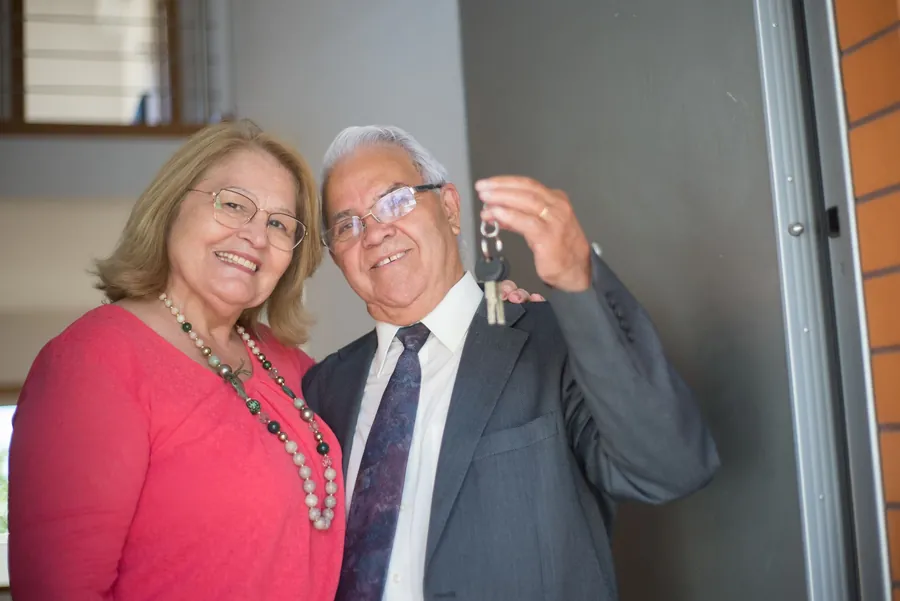Happy Hispanic-looking senior couple at a doorway holding up new house keys: woman in coral-pink top with beaded necklace, man in grey suit,