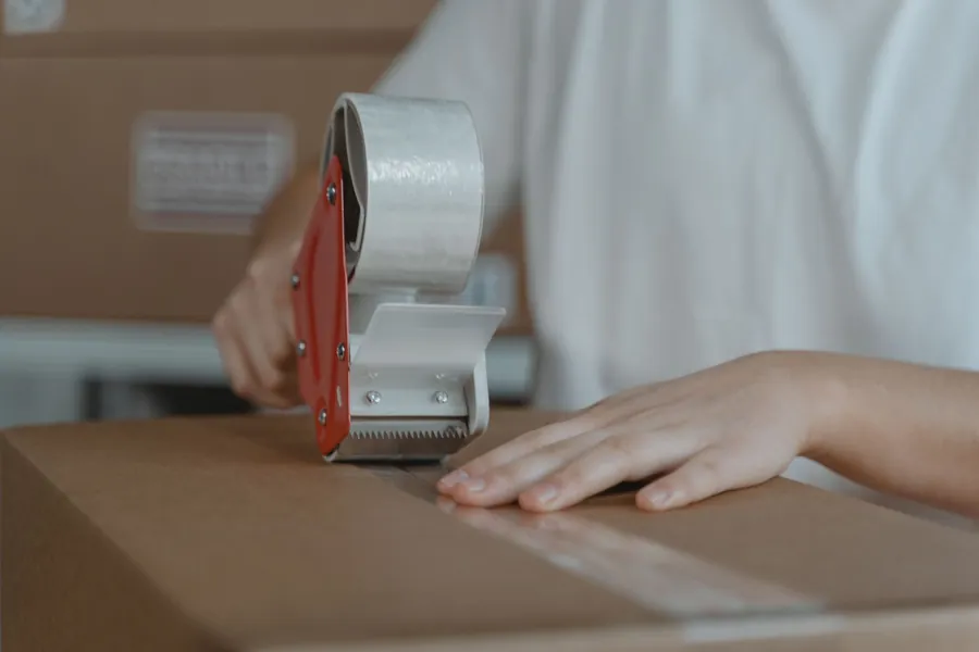 Close-up of hands running a red tape dispenser along the seam of a cardboard box
